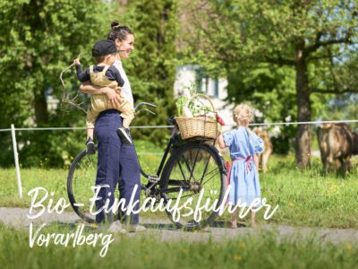 Junge Frau mit Kind auf dem Arm & Einkaufskorb und Fahrrad vor einer Weide