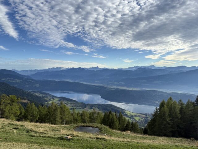 Landschaftsbild, von einer Anhöhe auf den Millstätter See, versteckt hinter Bäumen, darüber ein blauer Himmel mit Wolken